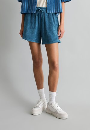 Osoba ubrana w niebieskie szorty z frotté, białe skarpetki, białe trampki i niebieską pasiastą kurtkę na jednolitym szarym tle.