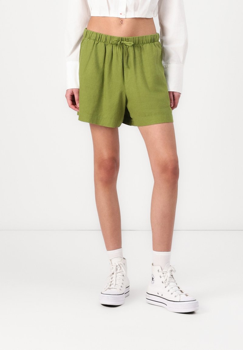 Vihreät puuvillashortsit, joissa on joustava vyötärönauha ja nyöritys; yhdistetty valkoisiin korkeavartisiin tennareihin ja valkoisiin sukkiin.