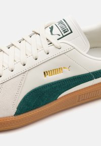 Sneakers bianchi con accenti in suede verde, parte superiore in pelle, lacci in tessuto e suola in gomma. Presentano un logo Puma dorato sul lato.
