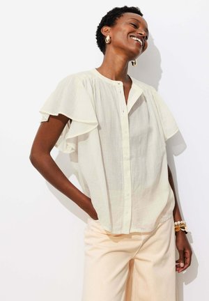Femme aux cheveux courts et bouclés, portant un chemisier blanc à manches courtes boutonné et un pantalon crème, souriante, une main dans la poche.