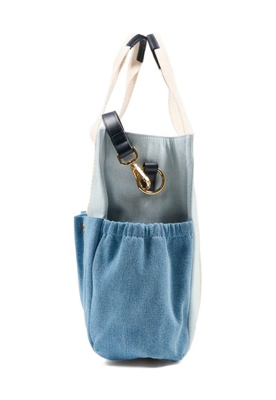 Lichtblauwe denim totebag met elastische zak aan de voorkant, zwarte leren bandaccenten en gouden metalen sluiting, hangend aan witte hengsels.