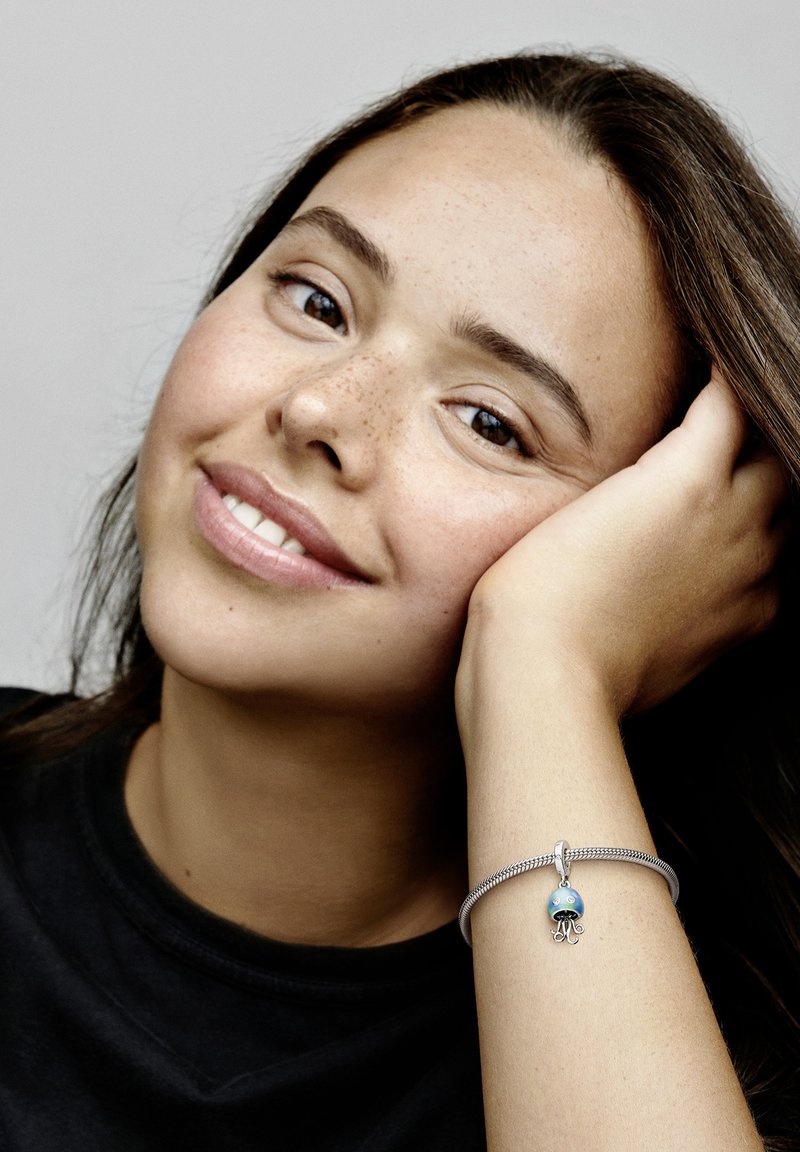 Srebrna zapestnica z modrim osličem, ki ima svetleče poudarke. Oseba, ki počiva s brado na roki, na enobarvni podlagi.