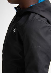 Schwarze Jacke mit Kapuze, mit einem blauen Innenfutter, strukturiertem Stoff und einem Logo-Patch auf der Vorderseite. Branding am Ärmel.