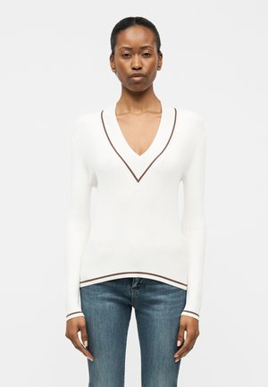 Mujer vistiendo un suéter de punto blanco con ribete marrón en el escote en V, puños y dobladillo, combinado con jeans de mezclilla azul, de pie frente a un fondo liso.