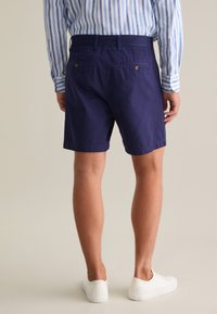 Marineblaue Shorts mit glatter Textur, ausgestattet mit zwei Gesäßtaschen mit Knopfverschlüssen und einem geraden Bein-Design, kombiniert mit weißen Sneakern.
