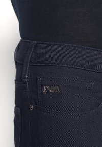 Mörkblå jeans med en texturerad tyg som har en liten metalllogotag och en enda bakficka. Synliga sömmar betonar detaljerna längs med sömmarna.