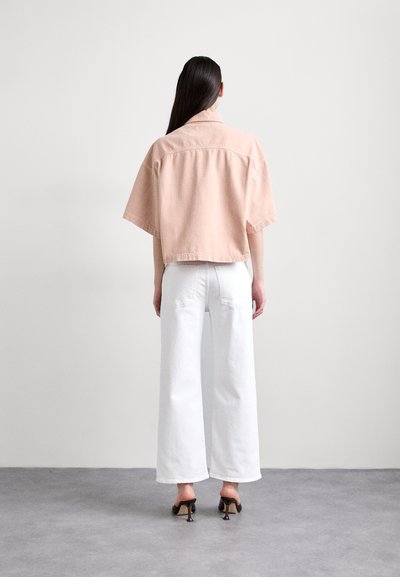Lichtroze, cropped denimjacket met korte mouwen, gecombineerd met witte, wijde broeken. Model staat tegen een neutrale achtergrond.