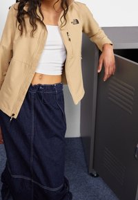 Beige zip-jacka med två sidofickor och en logotyp. Matchad med en ribbad vit crop top och en mörk denimkjol med dragsko i midjan.