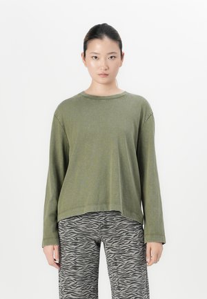 Jonge vrouw die recht vooruit kijkt, gekleed in een olijfgroen shirt met lange mouwen en grijze broek met zebra-print, tegen een witte achtergrond.