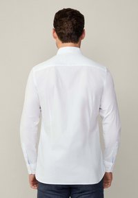 Camicia bianca a maniche lunghe con colletto classico, tessuto di cotone morbido e polsini abbottonati. Presenta un piccolo accento blu sulla manica.
