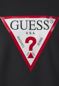 Logo triangulaire rouge et blanc avec "GUESS U.S.A." en haut, un grand point d'interrogation rouge au centre et les mots "WASHED JEANS" sur les côtés.