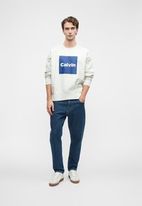 Mladý muž stojí s jednou rukou v vrecku, oblečený v béžovej mikine s modrým logom "Calvin", modrých džínsoch a bielych teniskách na bielom pozadí.