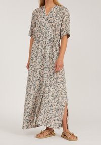 Femme portant une robe portefeuille florale beige à manches courtes, avec une fente sur le côté et des sandales en corde, sur un fond clair.