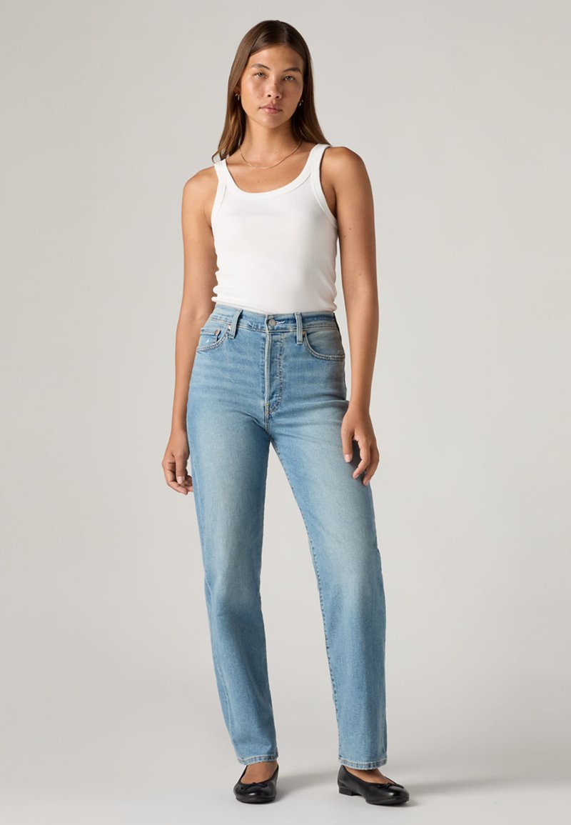 Jeans blu a vita alta con taglio a gamba dritta, abbinati a un top bianco aderente. Il tessuto appare morbido con una lavatura chiara e un leggero sbiadimento.