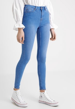 Vrouw draagt lichtblauwe skinny jeans, witte hoge sneakers en een witte blouse met lange mouwen en gerimpelde manchetten, staand tegen een effen achtergrond.