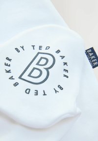 Witte stof met een cirkelvormig logo met de letter "B" in het midden. De tekst zegt "BAKER BY TED BAKER" met een label zichtbaar aan de zijkant.