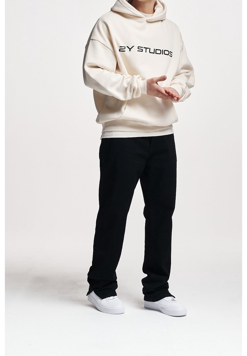 Personne portant un sweat à capuche crème avec le texte "2Y STUDIOS", un pantalon noir et des baskets blanches, debout avec les mains faisant un geste vers l'avant.