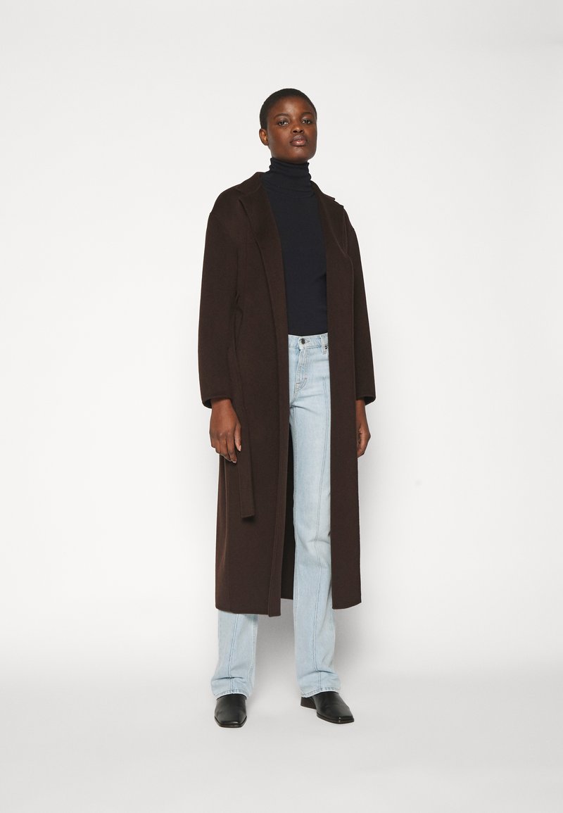 Lang brun frakke med sjalkrave, båret over en sort turtleneck, kombineret med lysblå jeans og sorte spidse sko.