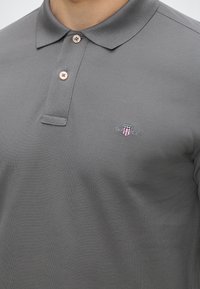 Polo gris avec deux boutons fermés et un petit logo écusson brodé en rouge, blanc et bleu sur le côté gauche de la poitrine.