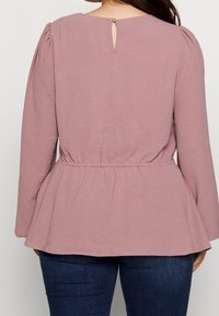 T-shirt à manches longues en tissu mauve doux et texturé, avec une taille cintrée, un style peplum et un petit détail en forme de serrure à l'arrière du col.