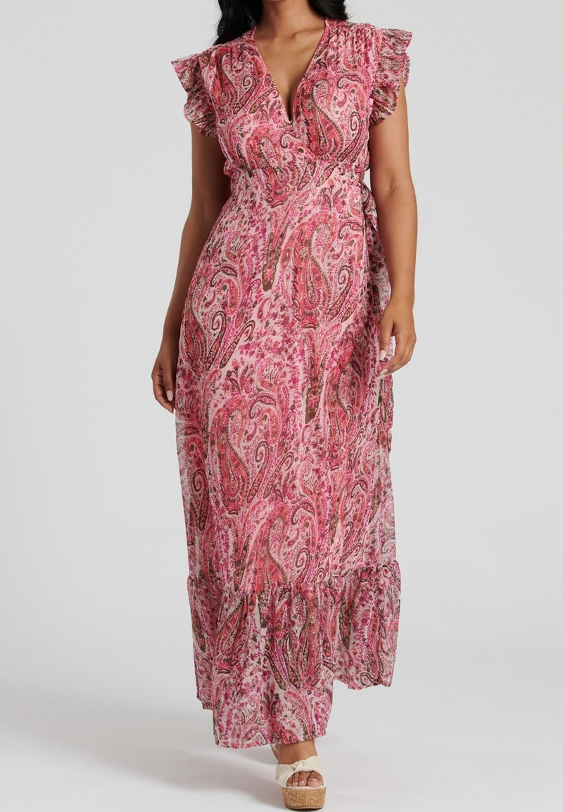 Femme portant une longue robe portefeuille rose à motif cachemire avec des manches courtes à volants et des sandales compensées beiges sur fond gris.