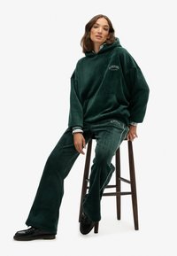 Grøn velour hoodie med en lomme foran, parret med matchende bukser med hvid piping og logo detaljer. Model siddende på en skammel.