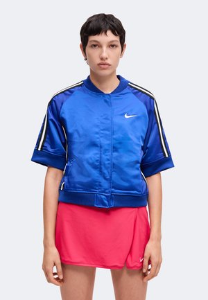 Osoba ubrana w niebieską kurtkę Nike z krótkim rękawem w czarno-białe paski oraz różową spódniczkę Nike na jednolitym tle.