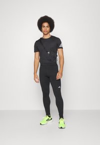 Maglietta atletica nera aderente, leggings neri e scarpe da ginnastica verde neon. Piccola borsa a tracolla nera con tracolla regolabile e dettaglio del logo.