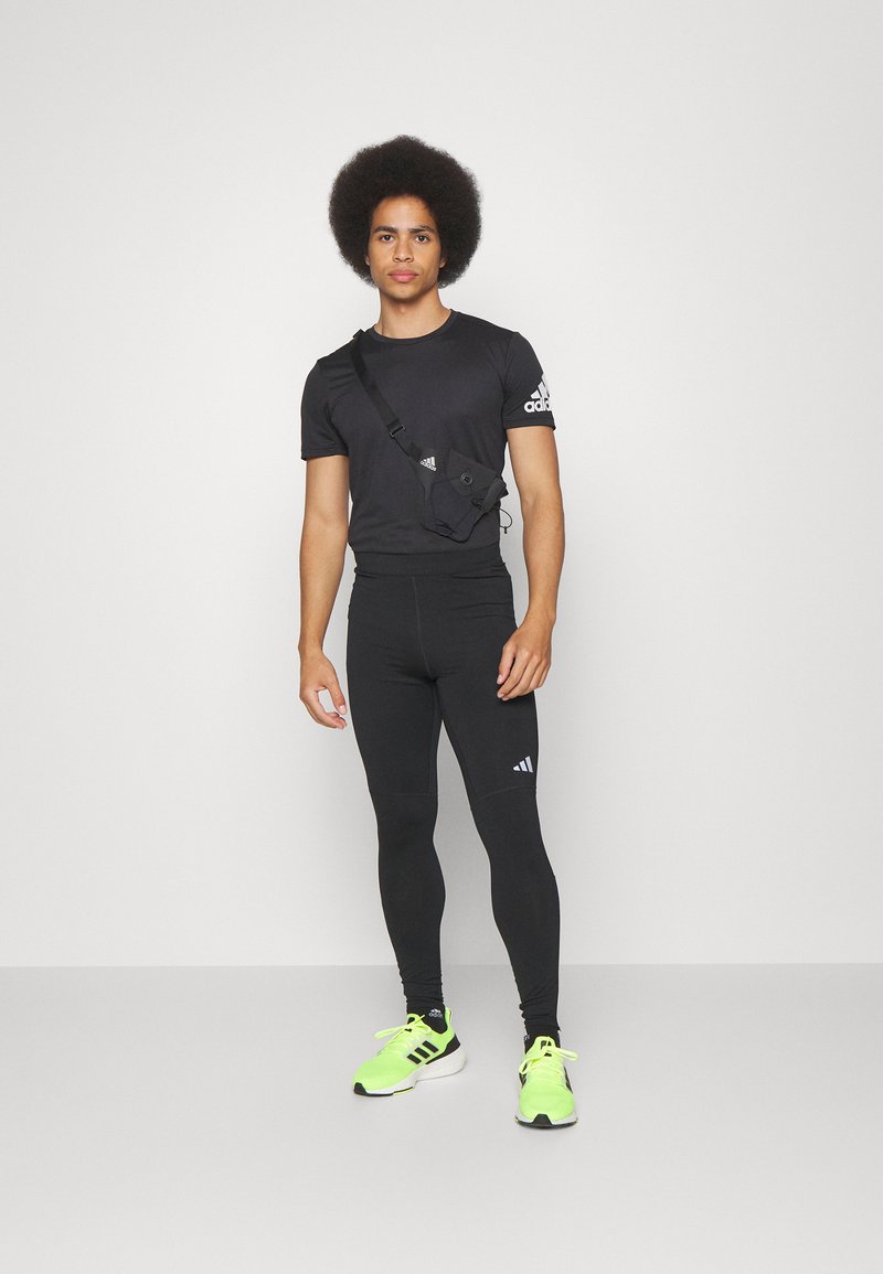 Zwarte sportieve fitted shirt, zwarte leggings en neon groene sneakers. Kleine zwarte schoudertas met verstelbare band en logo-detail.