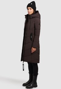 Brauner gesteppter Mantel mit Kapuze, diagonalem Steppmuster, seitlichen Taschen und Reißverschluss. Kombiniert mit schwarzen Leggings und Stiefeletten.