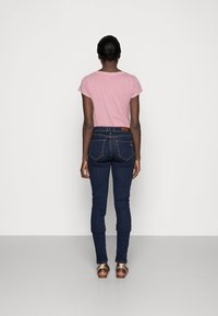 Camiseta rosa de manga corta, jeans de mezclilla oscuros de corte ajustado con bolsillos traseros y sandalias con hebilla de metal. Conjunto simple y casual visto desde la parte posterior.