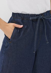 Marineblaue Hose mit elastischem Bund und Kordelzug, seitlichen Taschen und kontrastierenden Nähten. Hergestellt aus einem weichen Stoff mit leicht strukturierten Oberflächen.