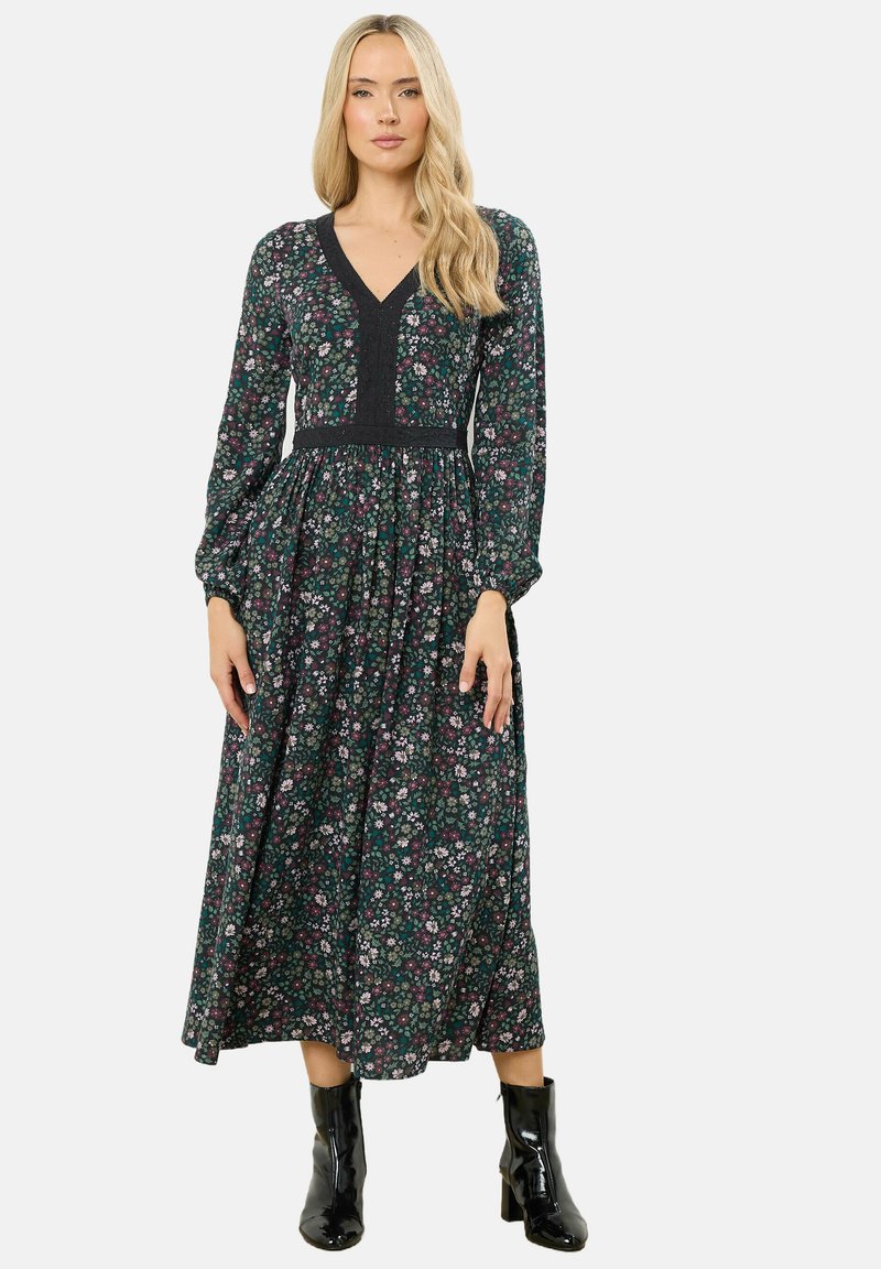 Vestido de manga larga y escote en V en un estampado floral verde oscuro. Presenta una cintura ajustada y una falda acampanada. Llevado con botas negras brillantes hasta el tobillo.