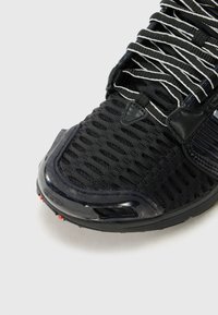 Chaussure de sport noire avec une tige en maille texturée, des lacets réfléchissants rayés et un embout en caoutchouc brillant avec des accents rouges.