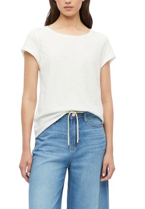 Vrouw draagt een effen witte T-shirt met korte mouwen, lichtjes ingestopt in lichtblauwe jeans met hoge taille en een witte tunnelsnoerceintuur.