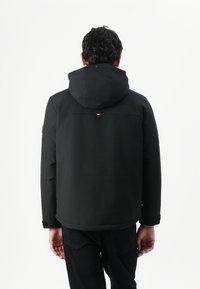 Sort hættejakke lavet af tekstureret stof, med et logovalg og justerbare manchetter. Viser bagsiden med minimalistisk design.