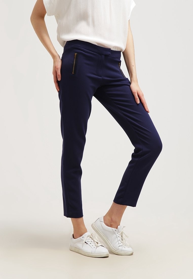 Marineblauwe slim-fit broek gemaakt van stretch stof, met gouden ritsaccenten op de voorzakken, gecombineerd met witte sneakers. Soepele textuur.