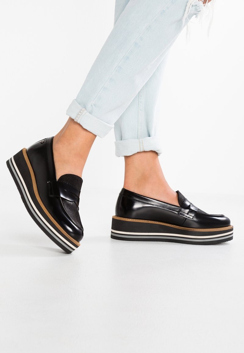 Personne portant un jean bleu clair retroussé et des mocassins plateforme noirs avec des semelles rayées, debout sur un sol blanc.