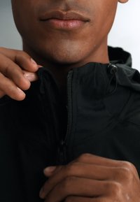 Le bas du visage et les mains d’une personne fermant une veste noire à col montant sur un fond uni.
