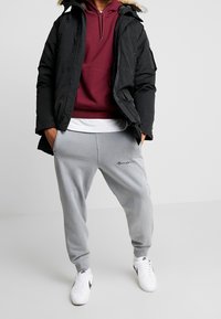Zwarte parka met bontkleurige rand, bordeauxrode hoodie, witte t-shirt en grijze joggers met borduursel, gecombineerd met witte sneakers.