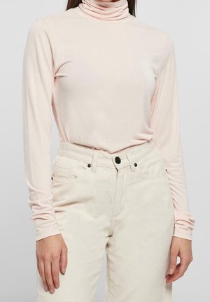 Femme portant un col roulé rose pâle à manches longues rentré dans un pantalon en velours côtelé écru taille haute avec un bouton noir et des rivets.