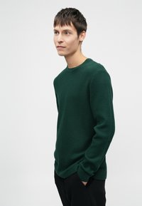 Σκούρο πράσινο πλεκτό πουλόβερ με στρογγυλή λαιμόκοψη και υφή. Επιτροπή μανικιών και χαλαρή εφαρμογή στο σώμα, συνδυασμένο με μαύρο παντελόνι.