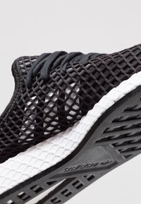 Svart idrottssko med övre i mesh, texturerade snören och en vit mellansula med grå detaljer. Gummisula med ett mönstrat mönster.