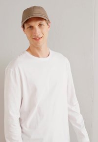 Weißes Langarmshirt mit Rundhalsausschnitt, kombiniert mit einer hellbraunen Kappe. Sanfte Textur, einfaches Design, neutrale Farben und entspannte Passform.