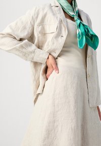 Chaqueta de lino beige con dos bolsillos en el pecho, superpuesta a una blusa beige clara y una falda de lino. Bufanda de seda verde con estampado atada al cuello.