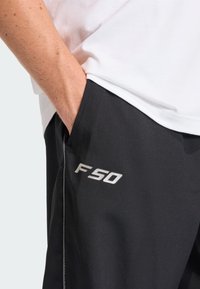 Svarte treningsshorts med sølvfarget "F50"-logo på venstre side, med glatt tekstur og sidelommer. Brukt sammen med en enkel hvit skjorte.