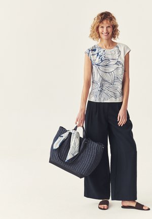 Žena sa usmieva, má na sebe biely top s kvetinovým vzorom a čierne široké nohavice, drží veľkú tmavomodrú pletenú tašku s vzorovaným šálom.