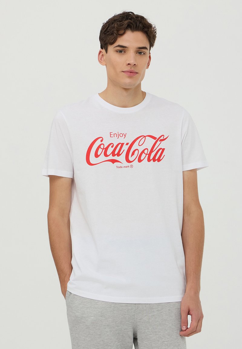 Maglietta bianca in cotone con la scritta "Enjoy Coca-Cola" in corsivo rosso. Vestibilità standard con maniche corte e collo a giro.