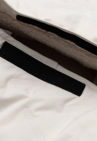 Witte kledingstuk met een gestructureerd oppervlak, zwart klittenbandsluiting en een zachte beige voering. Bevat subtiele patronen op het oppervlak.