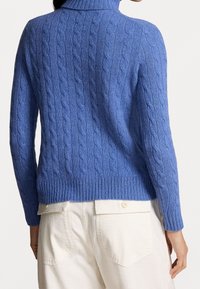 Kaapelikuvioinen sininen neulepaita, jossa on korkea kaulus ja resorireunus, yhdistetty vaaleanvärisiin rentoihin housuihin, joissa on takataskut.